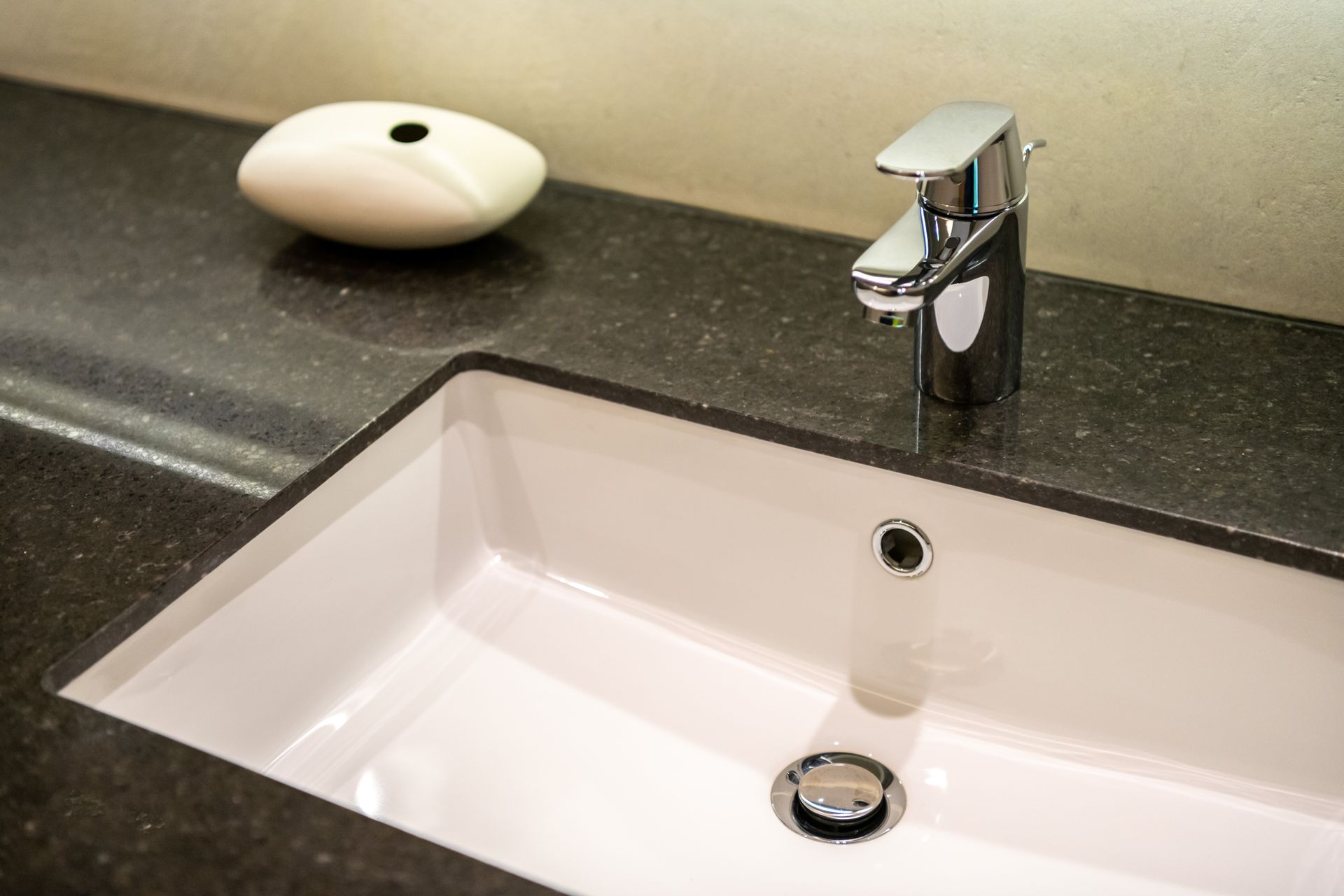 White bathroom sink with chrome faucet on a dark countertop and a small dish in the corner