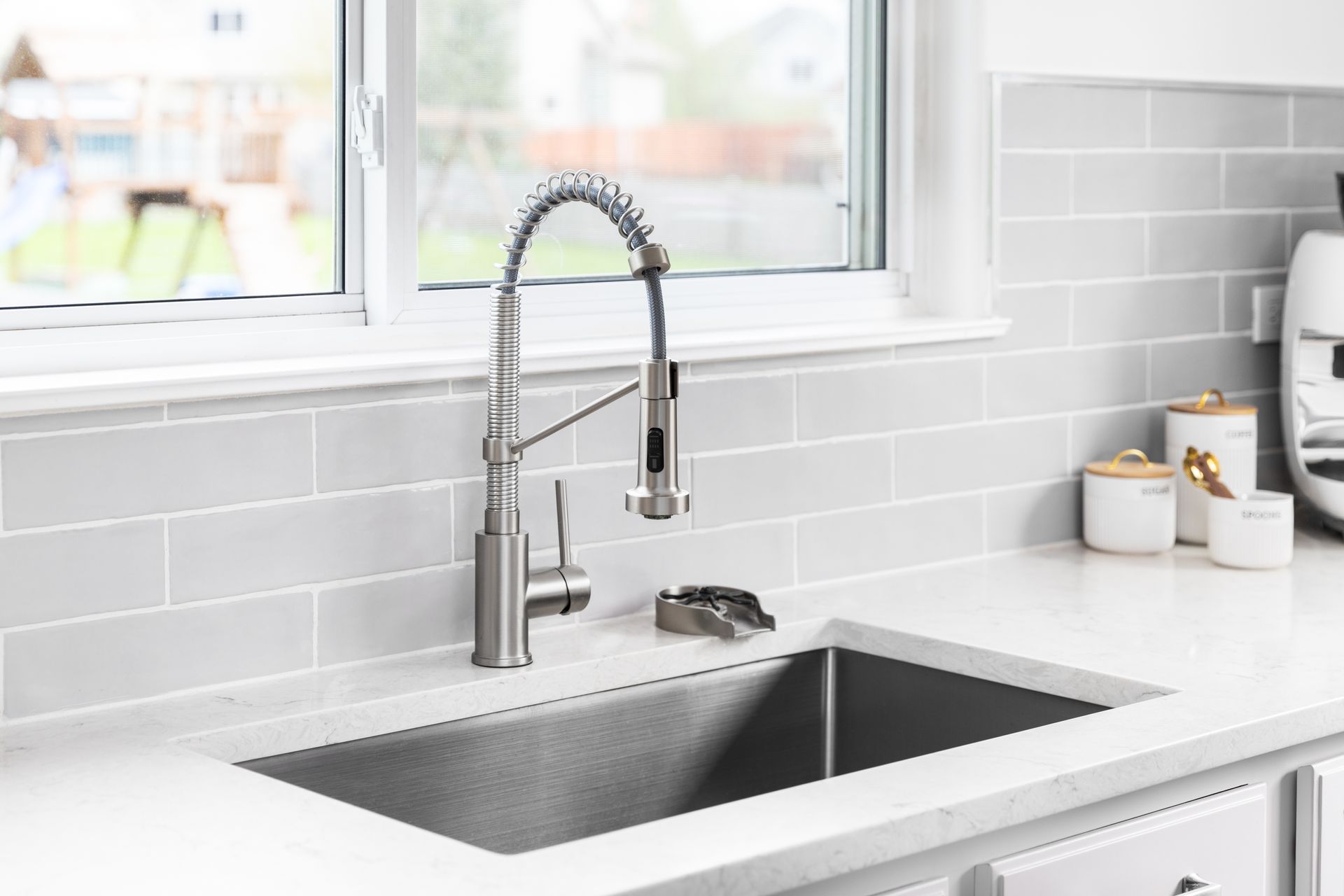 Modern kitchen sink with tall spring faucet, white tile backsplash, and bright window light