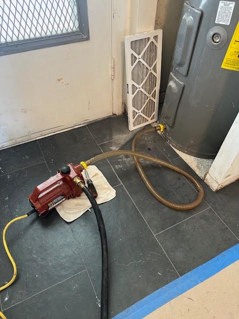 Red pressure test pump connected to hoses on a tiled floor beside a door and water heater.
