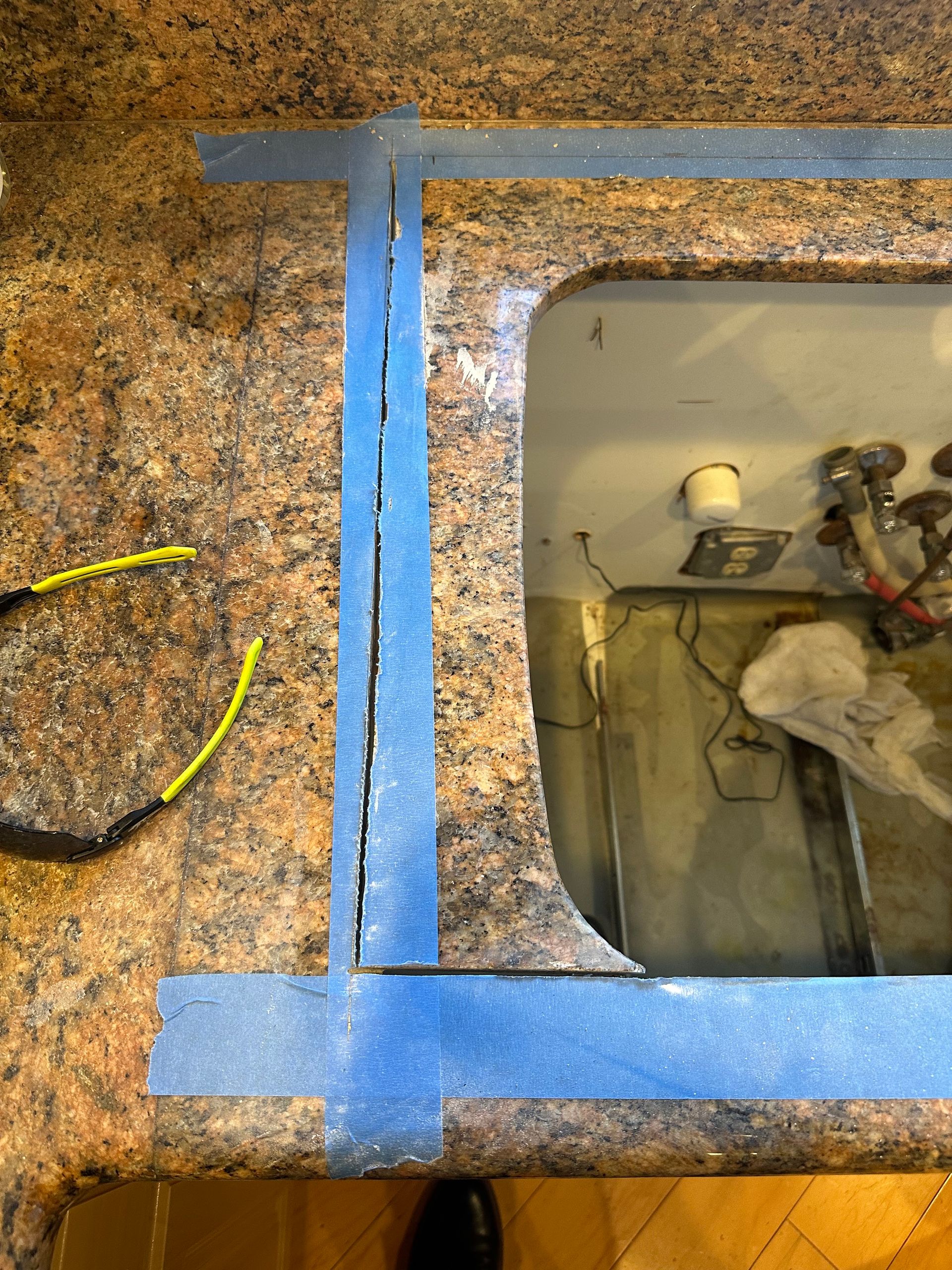 Blue-taped rectangular opening in a granite countertop, revealing plumbing and wires underneath