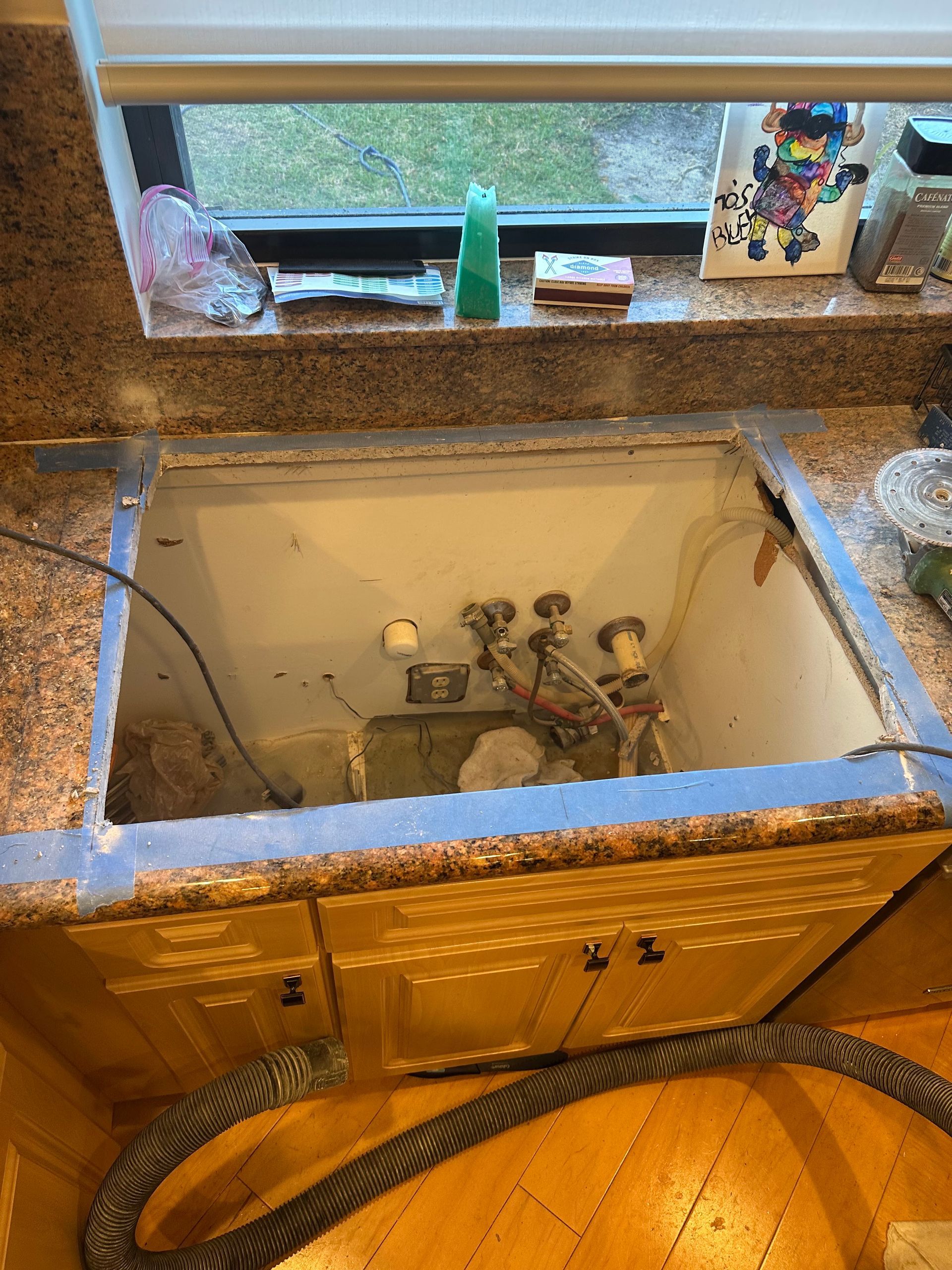 Kitchen sink cabinet under a countertop, with exposed plumbing and a hose on the floor beneath the open sink basin.