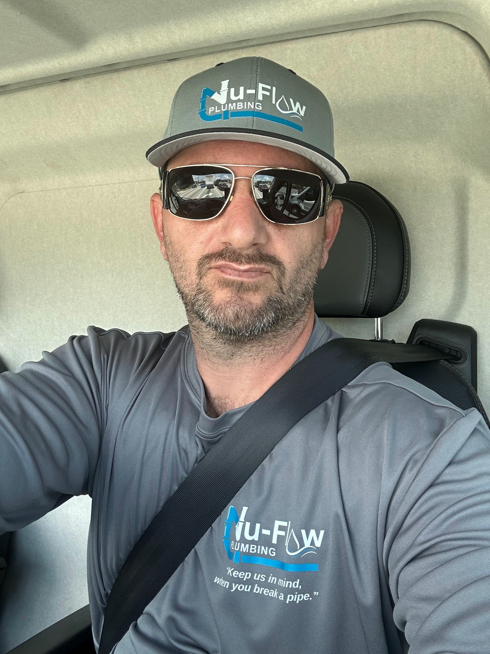 Man in a gray shirt and cap with sunglasses, seated in a vehicle, wearing a seatbelt and a company logo.
