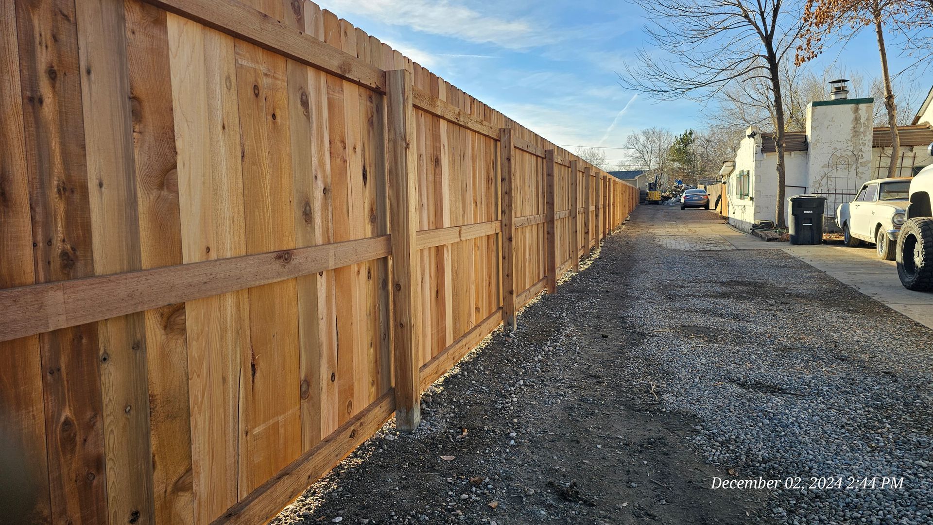 A newly installed wooden fence runs along a gravel path next to residential buildings under a clear blue sky.
