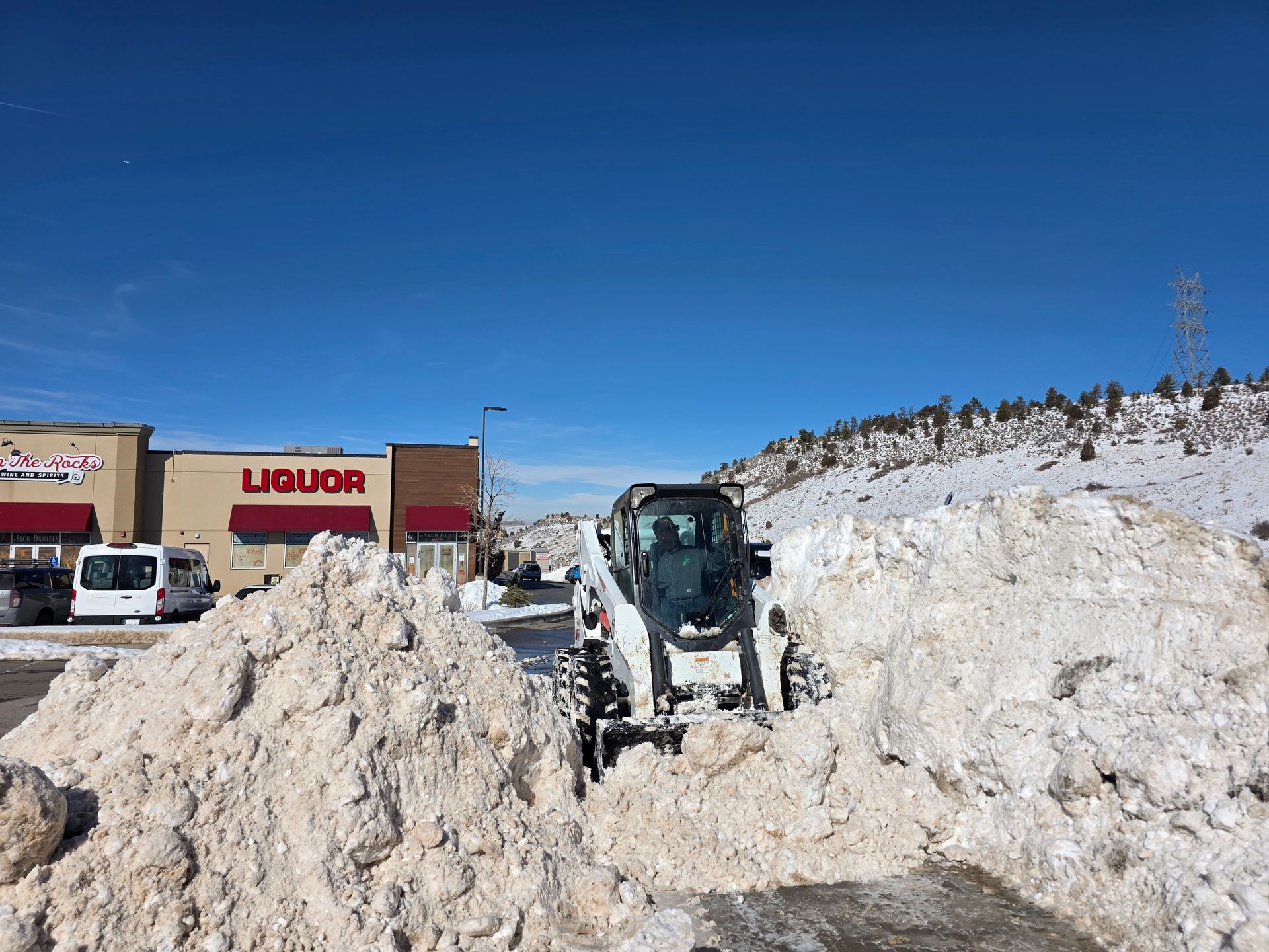 A white skid steer loader clears deep snow in a commercial parking lot near a strip mall under a clear blue sky.