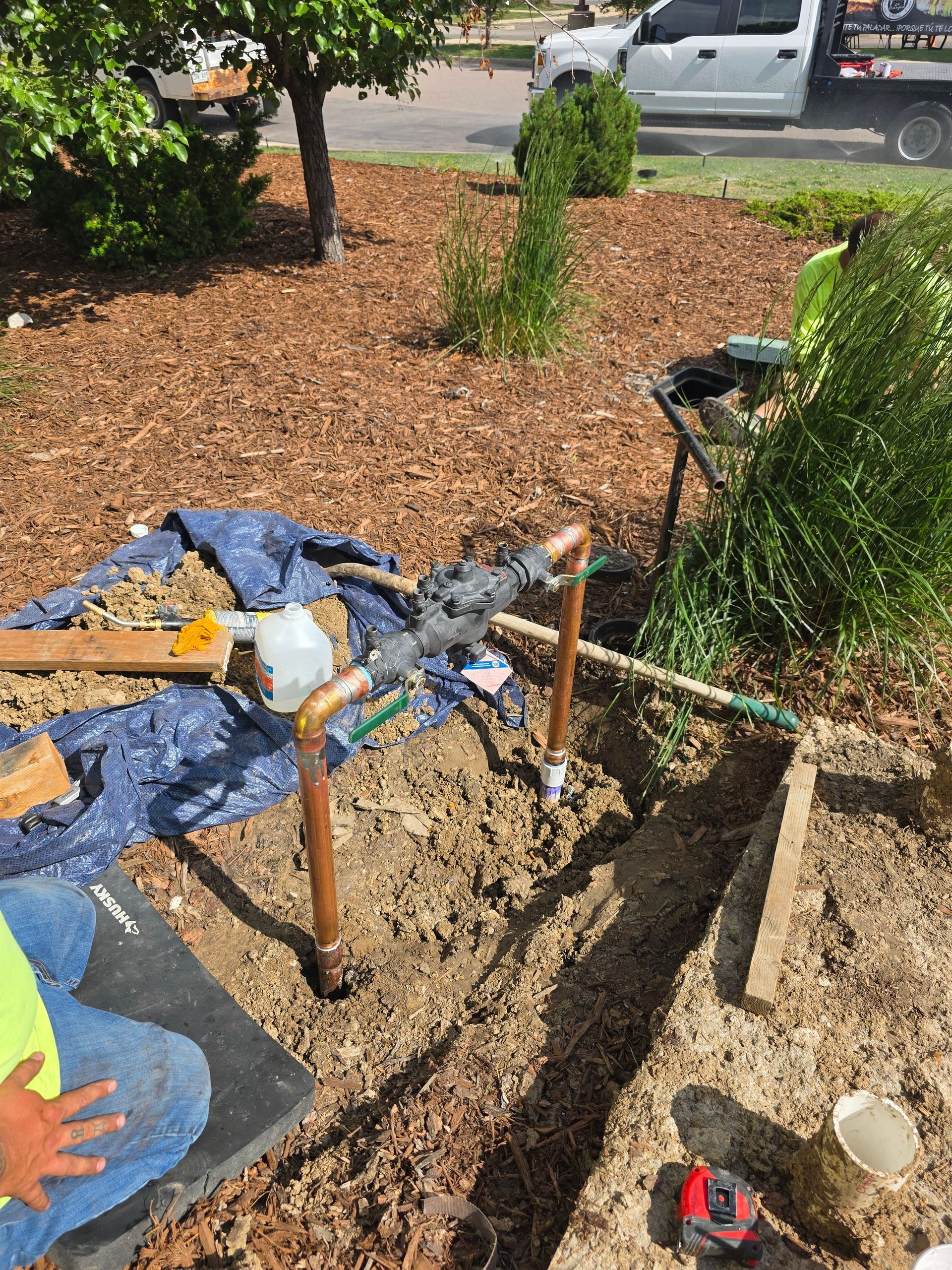 Plumbing repair with copper pipes in a shallow trench in a yard, with a person visible nearby and a white truck in back.