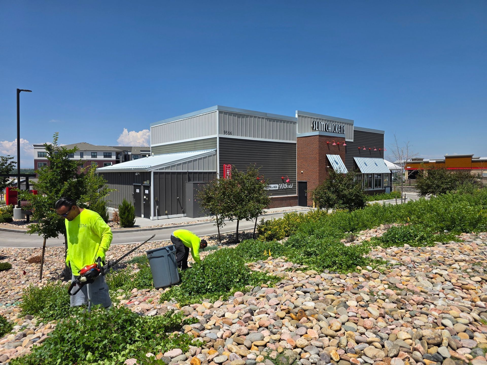 Two workers in high-visibility neon yellow shirts trim weeds near a rocky landscaping bed in front of a commercial building.