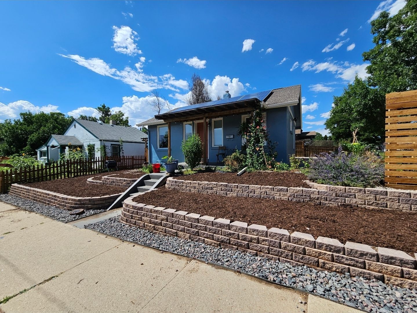A blue house with a solar-paneled roof, tiered landscaping, and stone retaining walls under a bright, partly cloudy sky.