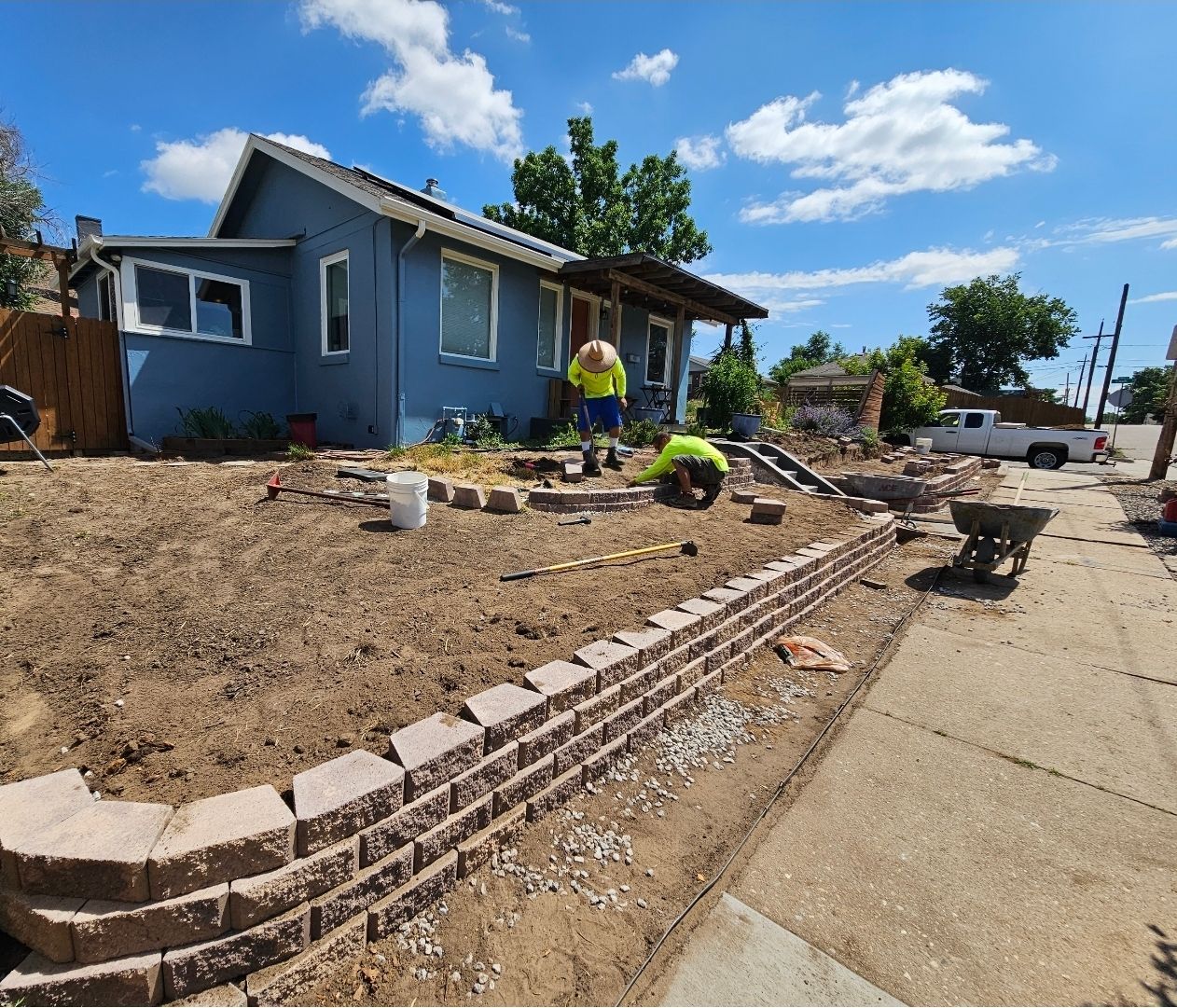 Two workers in bright vests build a retaining wall in front of a blue house on a sunny day.