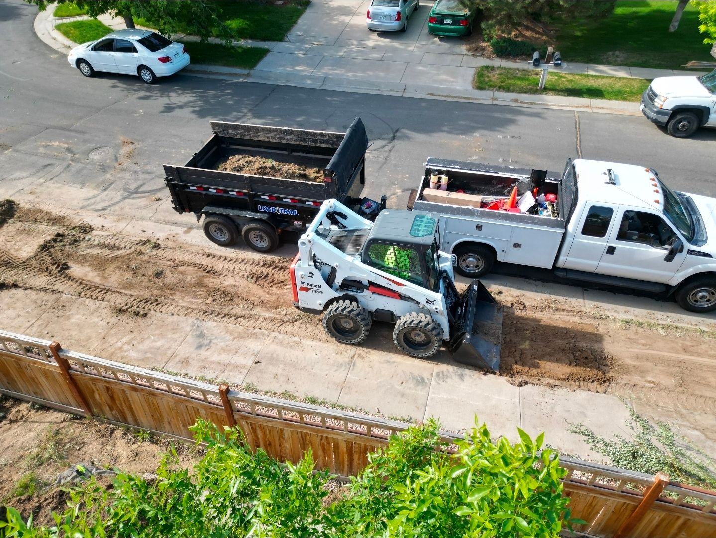 A white skid-steer loader works on a dirt patch near a white truck and a dump trailer parked on a residential street.