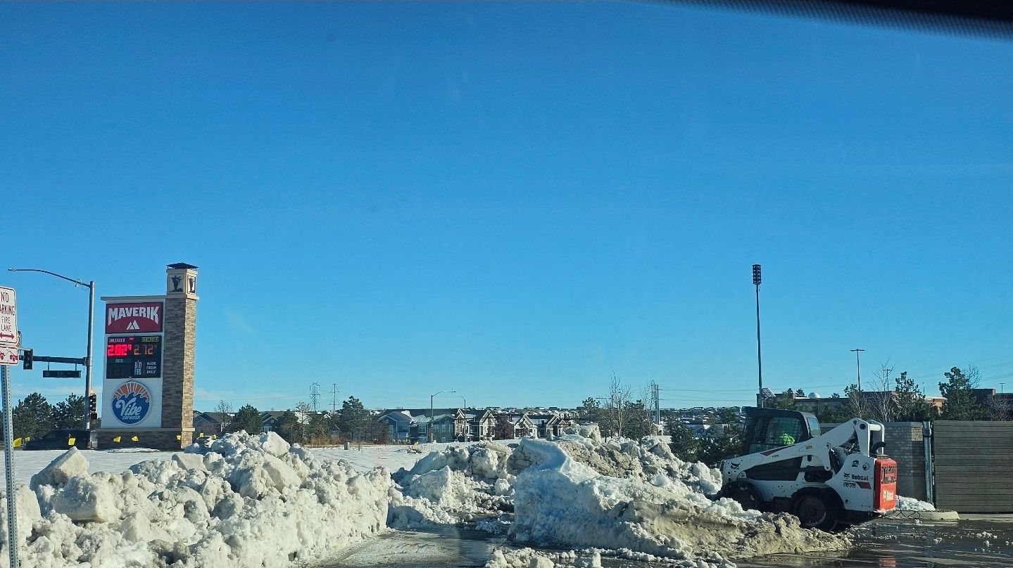 A white skid-steer loader clears snow near a tall, stone-based sign under a clear blue sky.