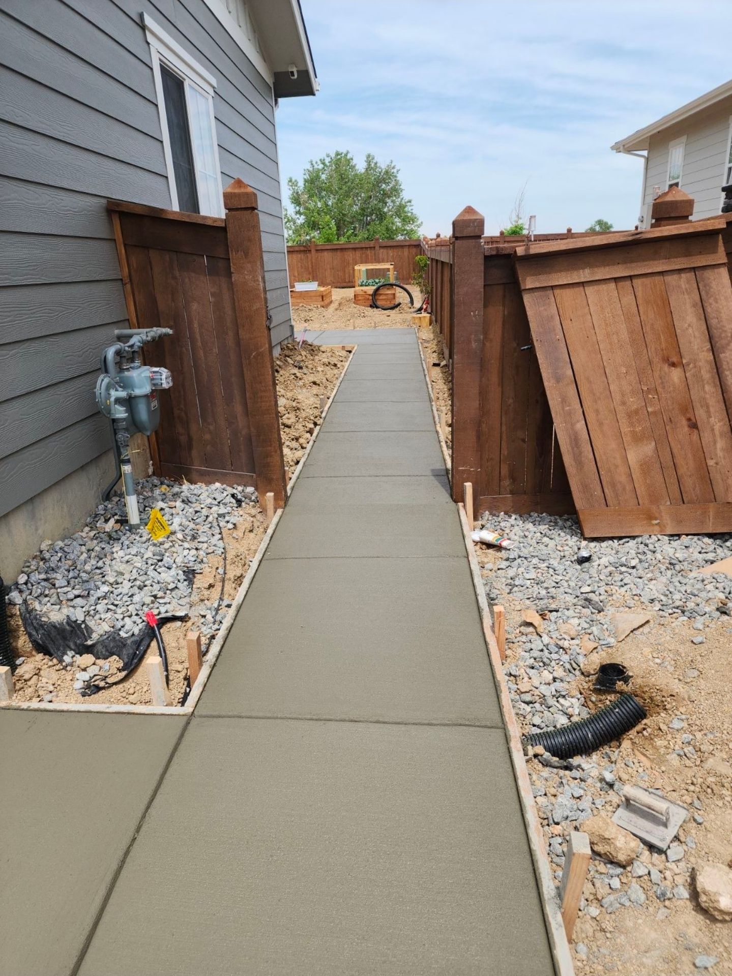 A newly poured concrete walkway runs between two wooden fences along the side of a grey house with gravel ground covering.