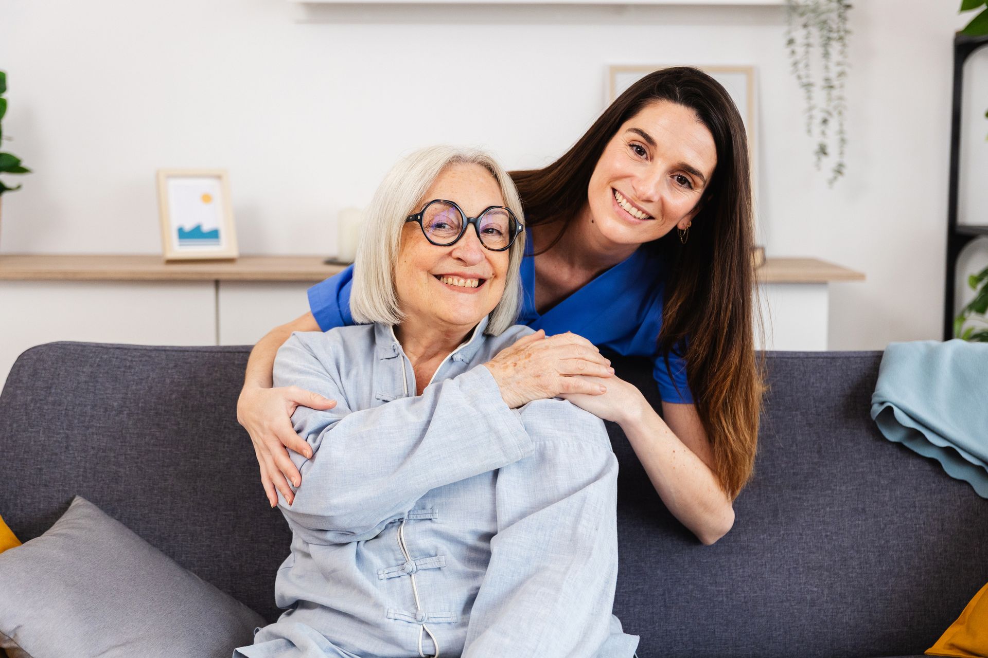Woman hugs smiling older person sitting on a sofa. Both looking at the camera. Woman hugs smiling older person sitting on a sofa. Both looking at the camera.