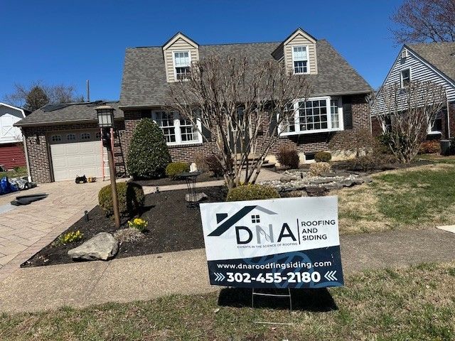 A lawn sign for DNA Roofing and Siding is in front of a brick house with dormer windows and a landscaped yard.