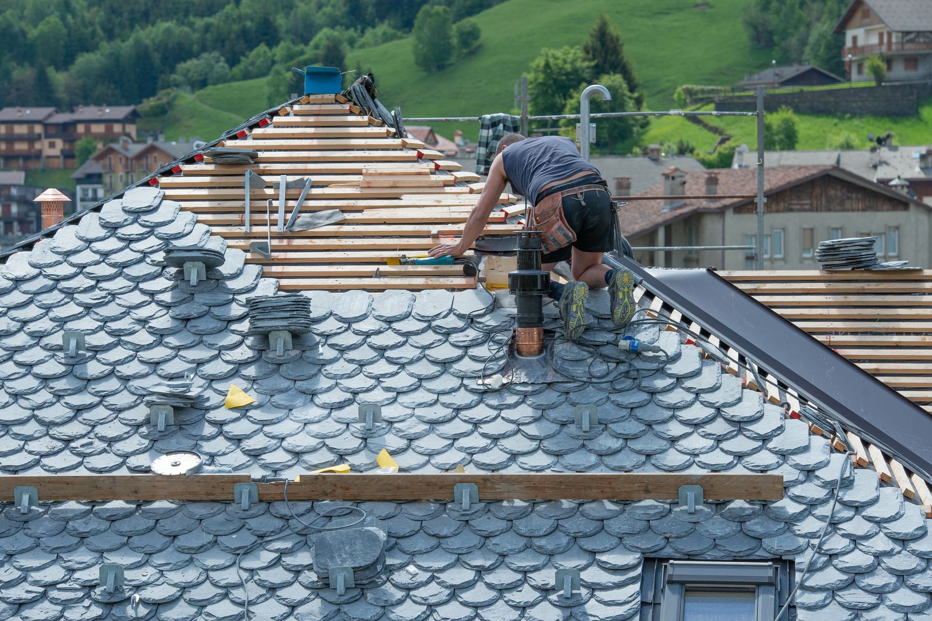 Roofer on a roof installing gray scalloped shingles, with a backdrop of a green hillside and buildings. Roofer on a roof installing gray scalloped shingles, with a backdrop of a green hillside and buildings.