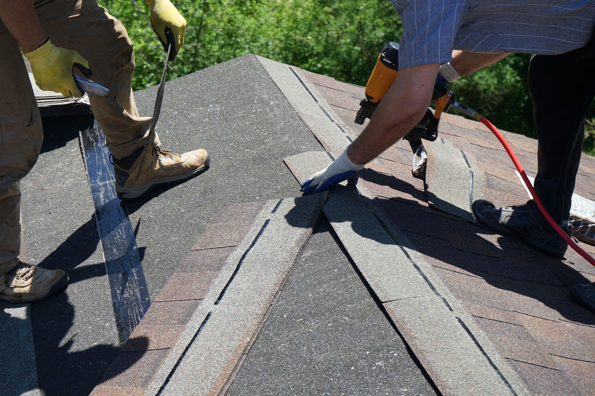 Roofers installing shingles on a roof peak using a nail gun; sunny outdoor setting. Roofers installing shingles on a roof peak using a nail gun; sunny outdoor setting.