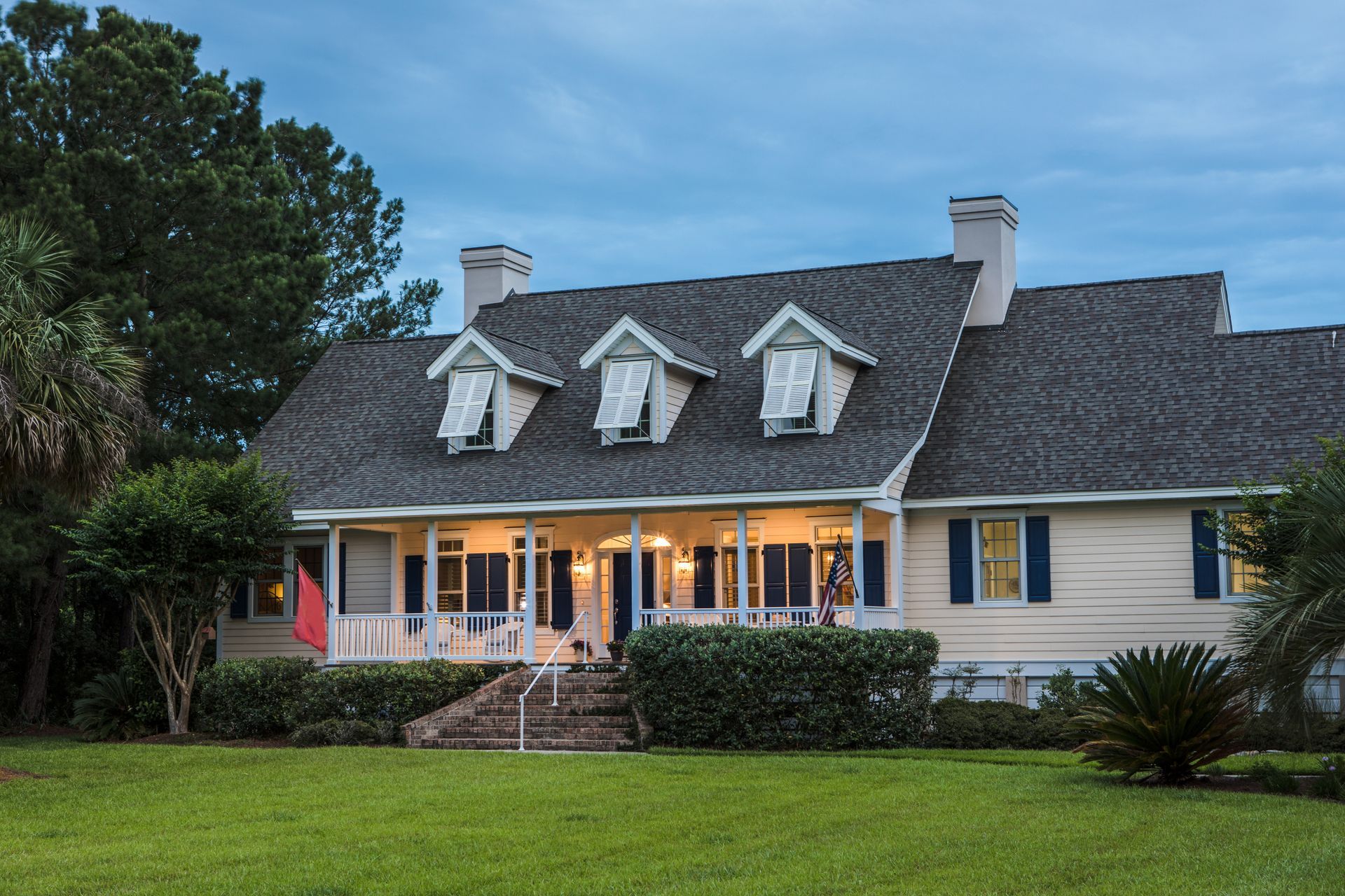Cottage-style home with three dormers, porch, and shutters, on a lawn with trees in the background. Cottage-style home with three dormers, porch, and shutters, on a lawn with trees in the background.