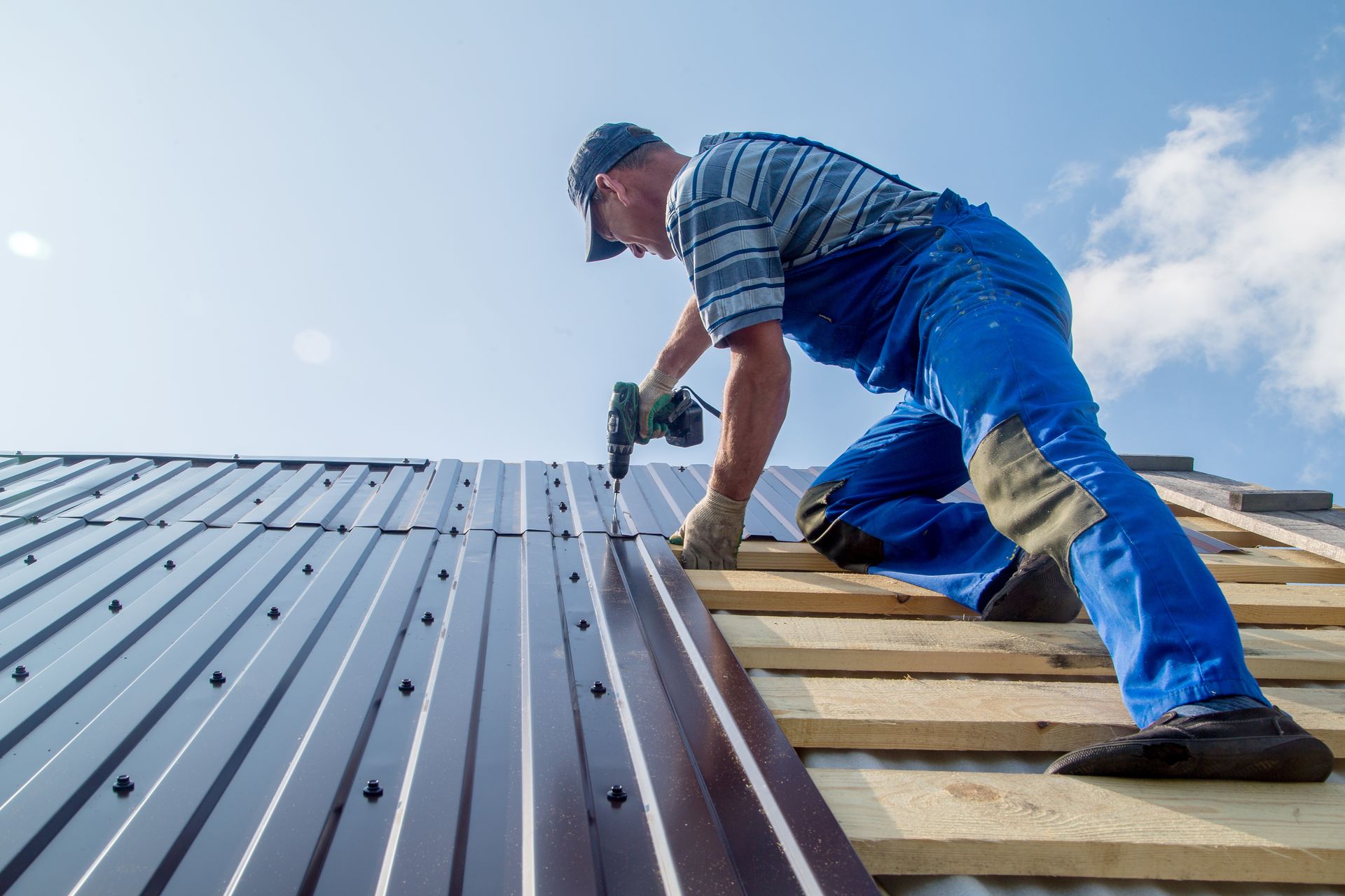 A roofer in blue overalls and cap installing metal roofing on a sunny day. A roofer in blue overalls and cap installing metal roofing on a sunny day.