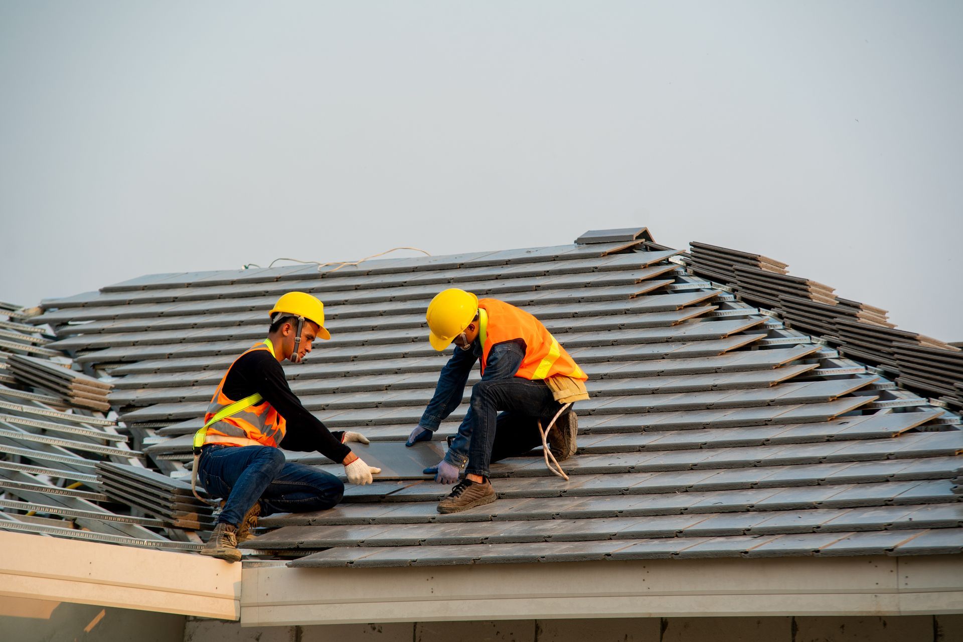 Two roofers in hard hats and vests install gray tiles on a roof. Two roofers in hard hats and vests install gray tiles on a roof.