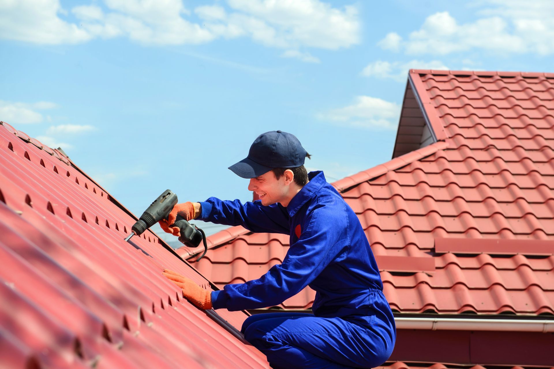 Roofer in blue jumpsuit and cap uses drill on red tile roof against blue sky. Roofer in blue jumpsuit and cap uses drill on red tile roof against blue sky.