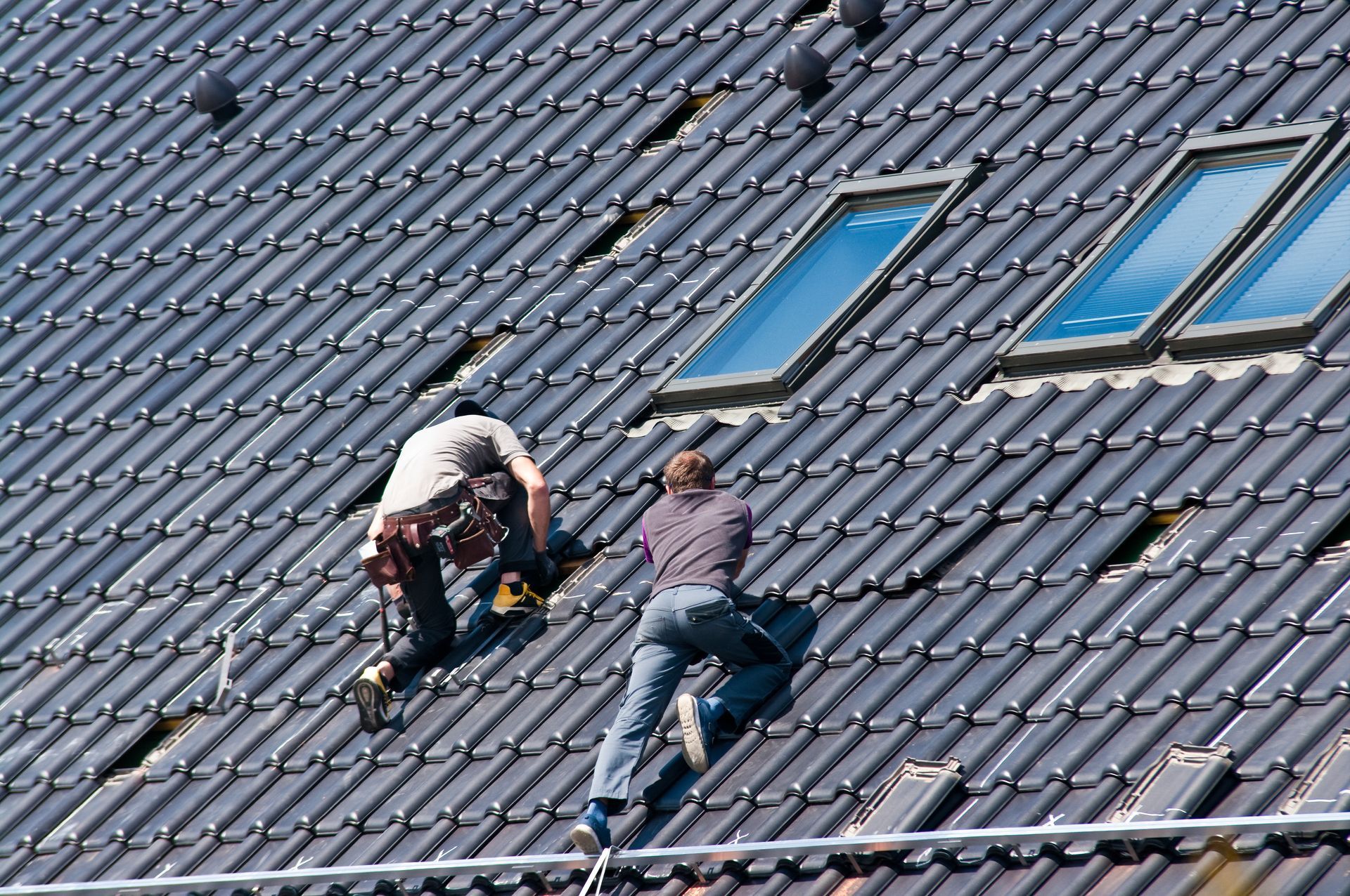 Two roofers on a tiled roof near skylights; one uses a tool. Two roofers on a tiled roof near skylights; one uses a tool.