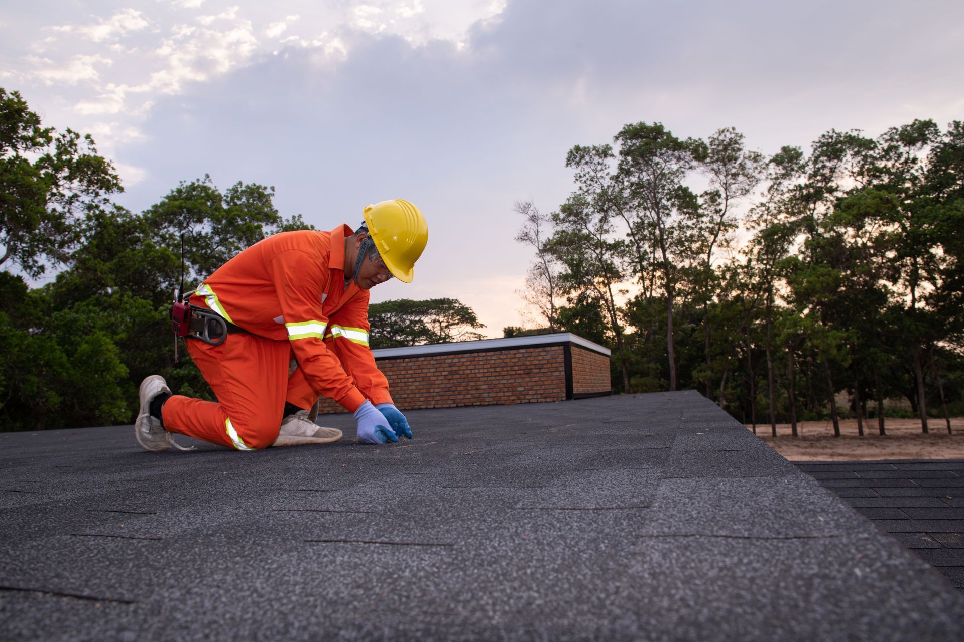 Roofer kneels on a dark shingled roof, wearing orange overalls and a yellow hard hat. Trees and cloudy sky in the background. Roofer kneels on a dark shingled roof, wearing orange overalls and a yellow hard hat. Trees and cloudy sky in the background.