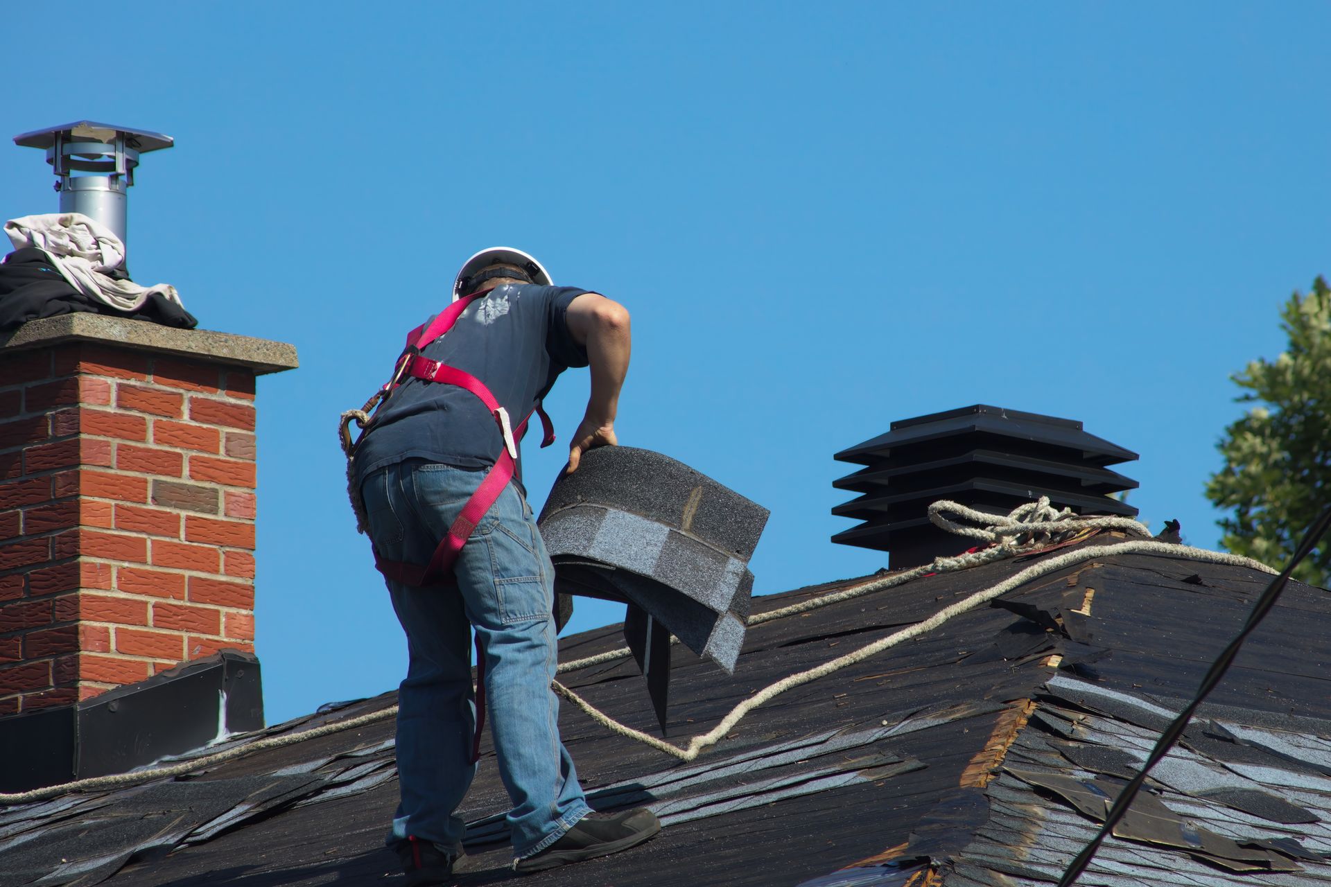Roofer on a dark roof replacing shingles, wearing a safety harness near a brick chimney. Roofer on a dark roof replacing shingles, wearing a safety harness near a brick chimney.