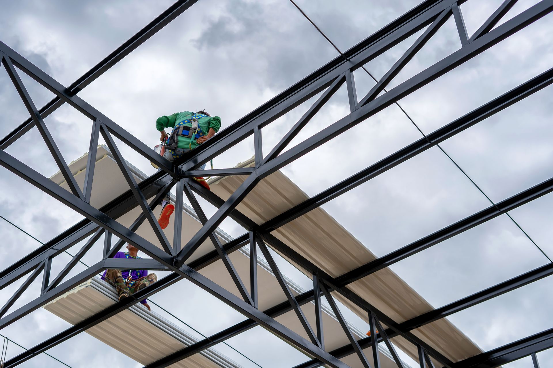 Construction workers installing roofing panels on a steel beam structure under a cloudy sky. Construction workers installing roofing panels on a steel beam structure under a cloudy sky.