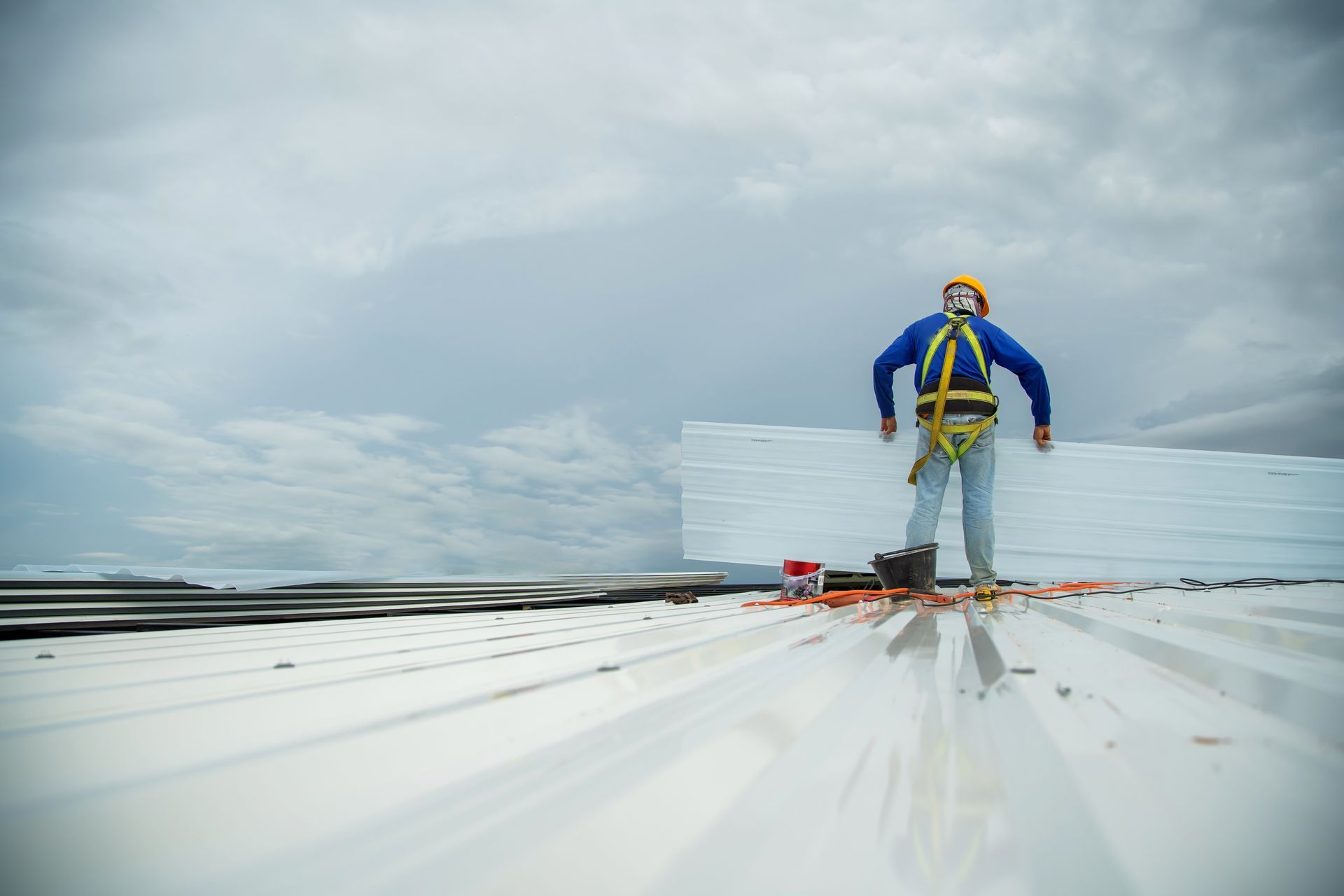 Roofer in safety harness on a white metal roof, holding foam insulation against a cloudy sky. Roofer in safety harness on a white metal roof, holding foam insulation against a cloudy sky.