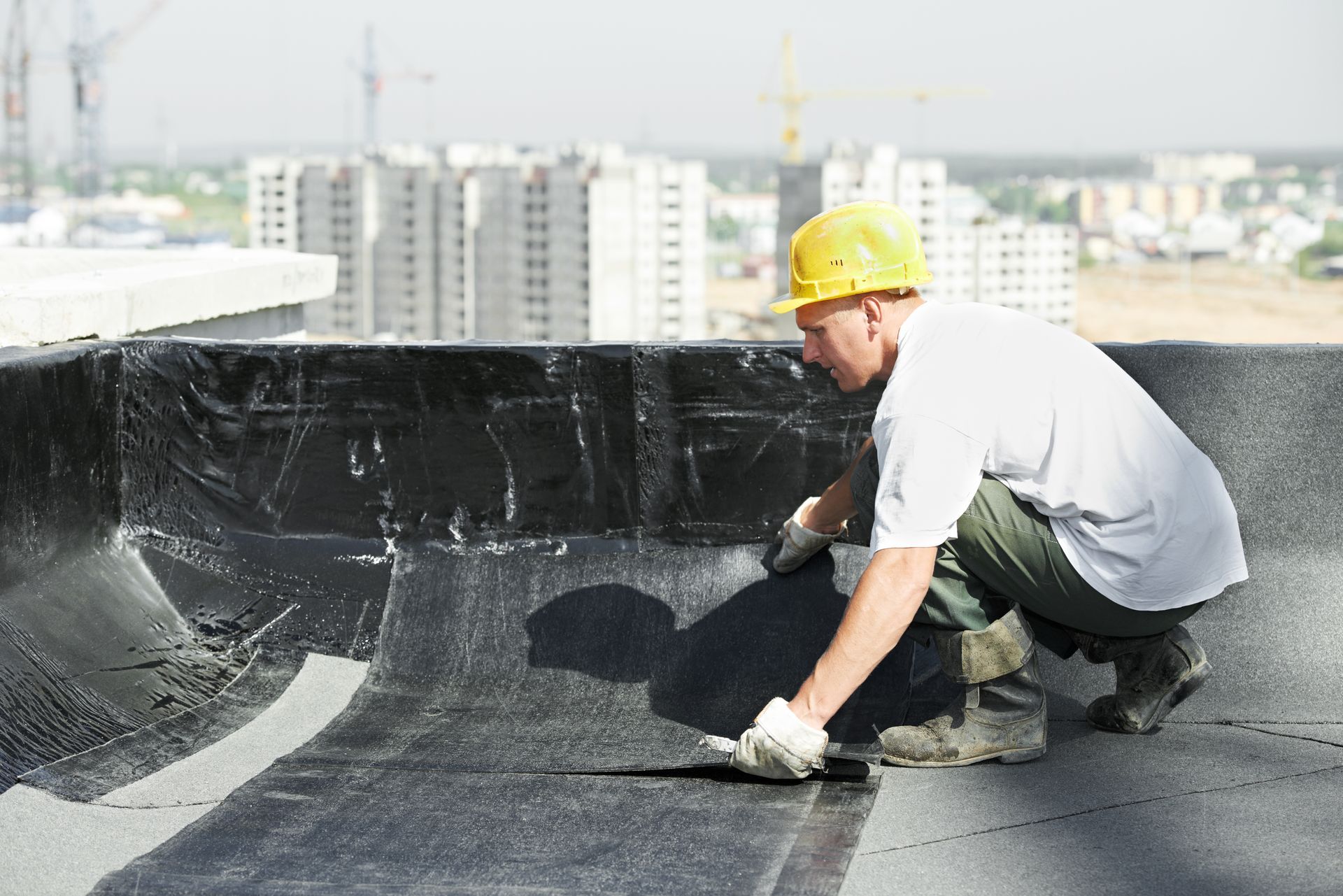 Roofer in hard hat on flat roof, applying black sealant. City buildings in the background. Roofer in hard hat on flat roof, applying black sealant. City buildings in the background.