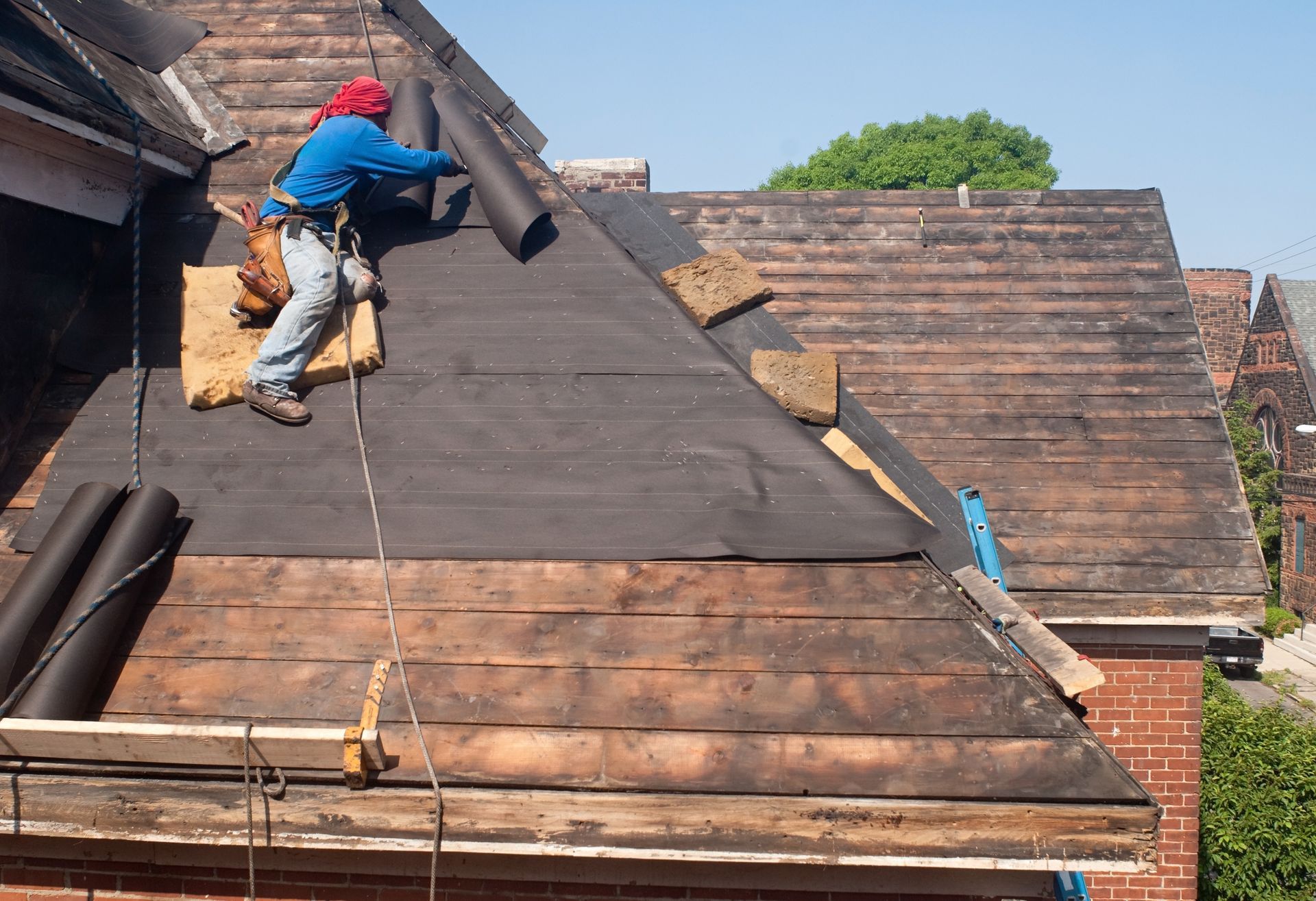 Roofer on a roof, installing black underlayment. Brick building, blue sky. Roofer on a roof, installing black underlayment. Brick building, blue sky.