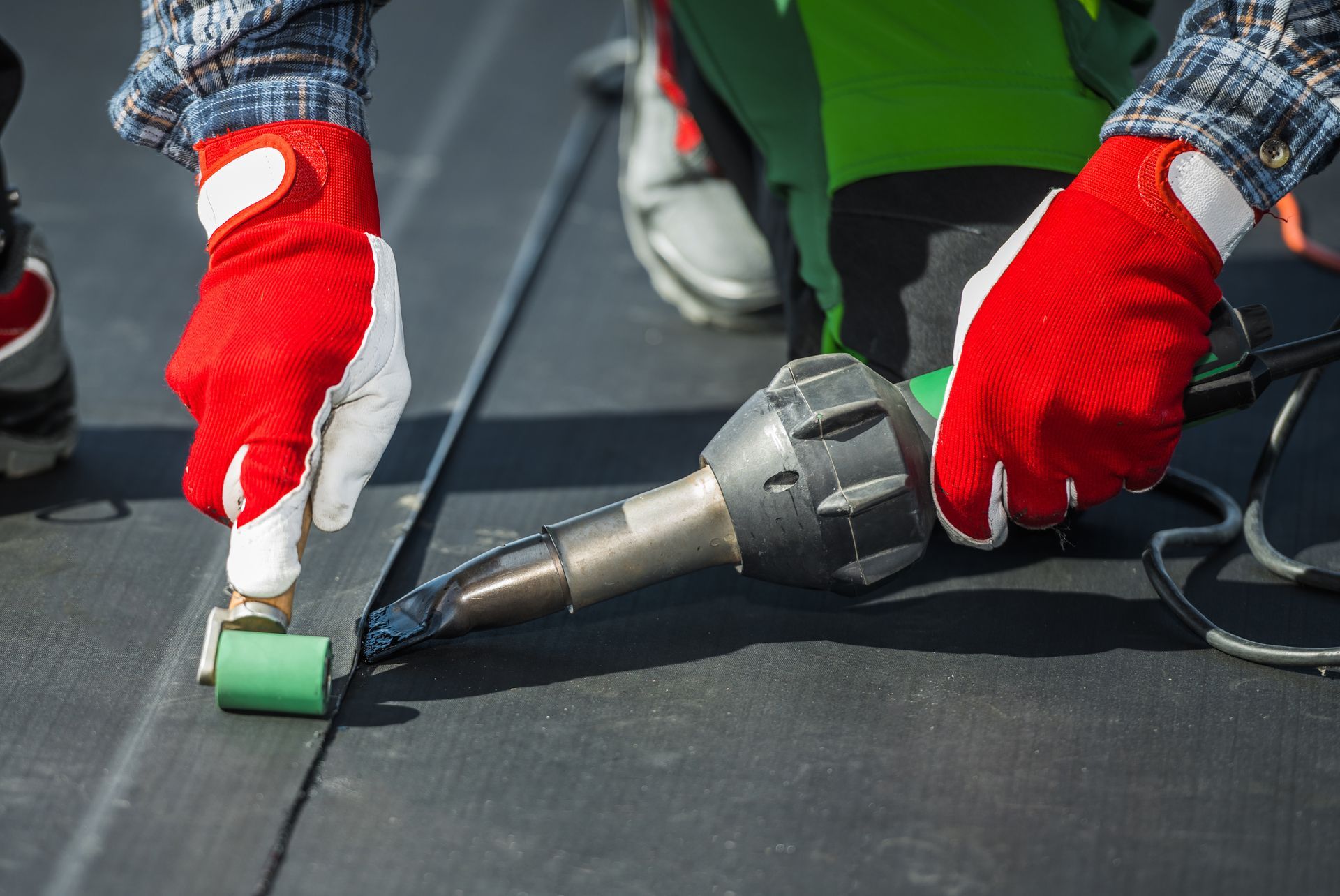 Roofer using a heat gun and roller to seal a black roof seam. Red and white gloves. Roofer using a heat gun and roller to seal a black roof seam. Red and white gloves.
