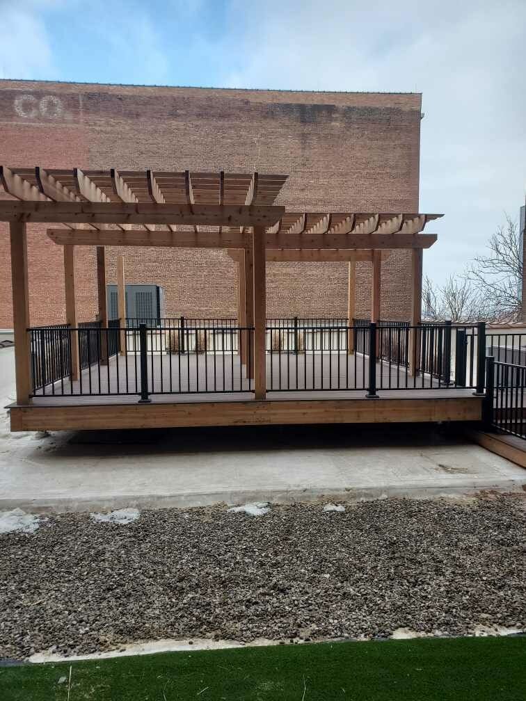 A wooden deck with a pergola and railing in front of a brick building.