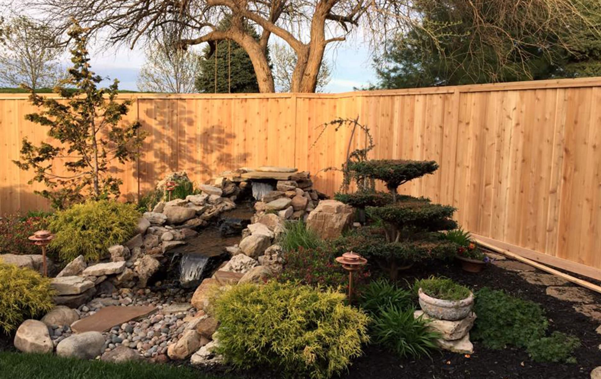 A wooden fence surrounds a garden with a waterfall and rocks.