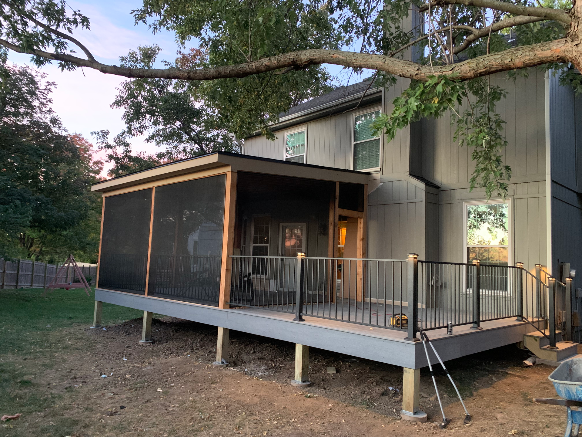 A house with a grey deck, screened porch, and black railing. Backyard setting, under a tree.