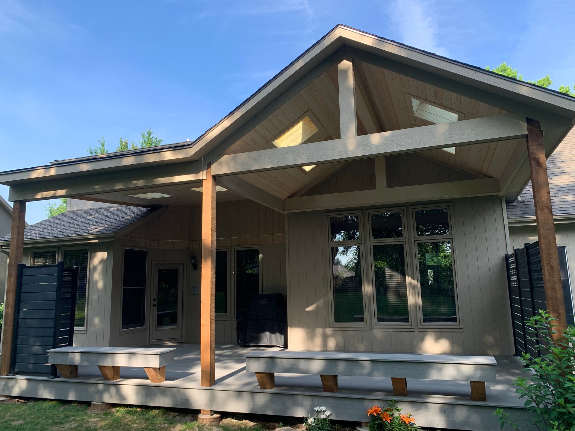 Tan covered patio attached to a house with wooden beams, bench seating, and windows.