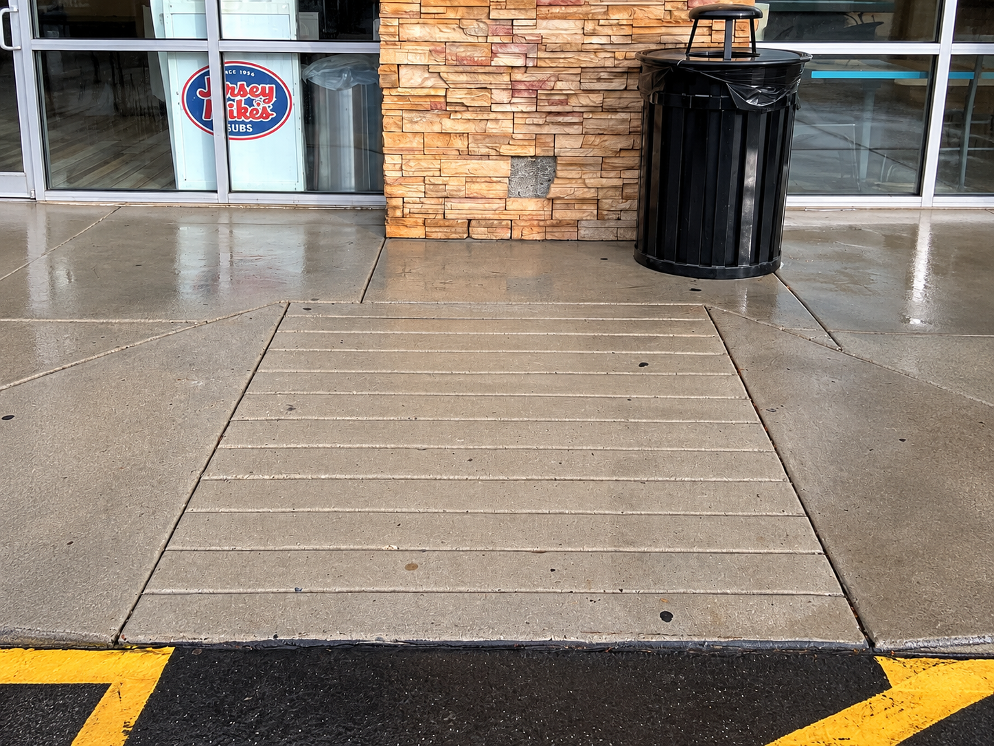 Concrete wheelchair ramp leading to a storefront entrance beside a black trash can.