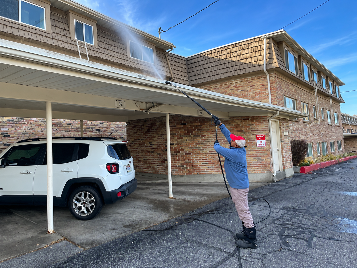 Person pressure washing a brick apartment building beside a white SUV under a carport.