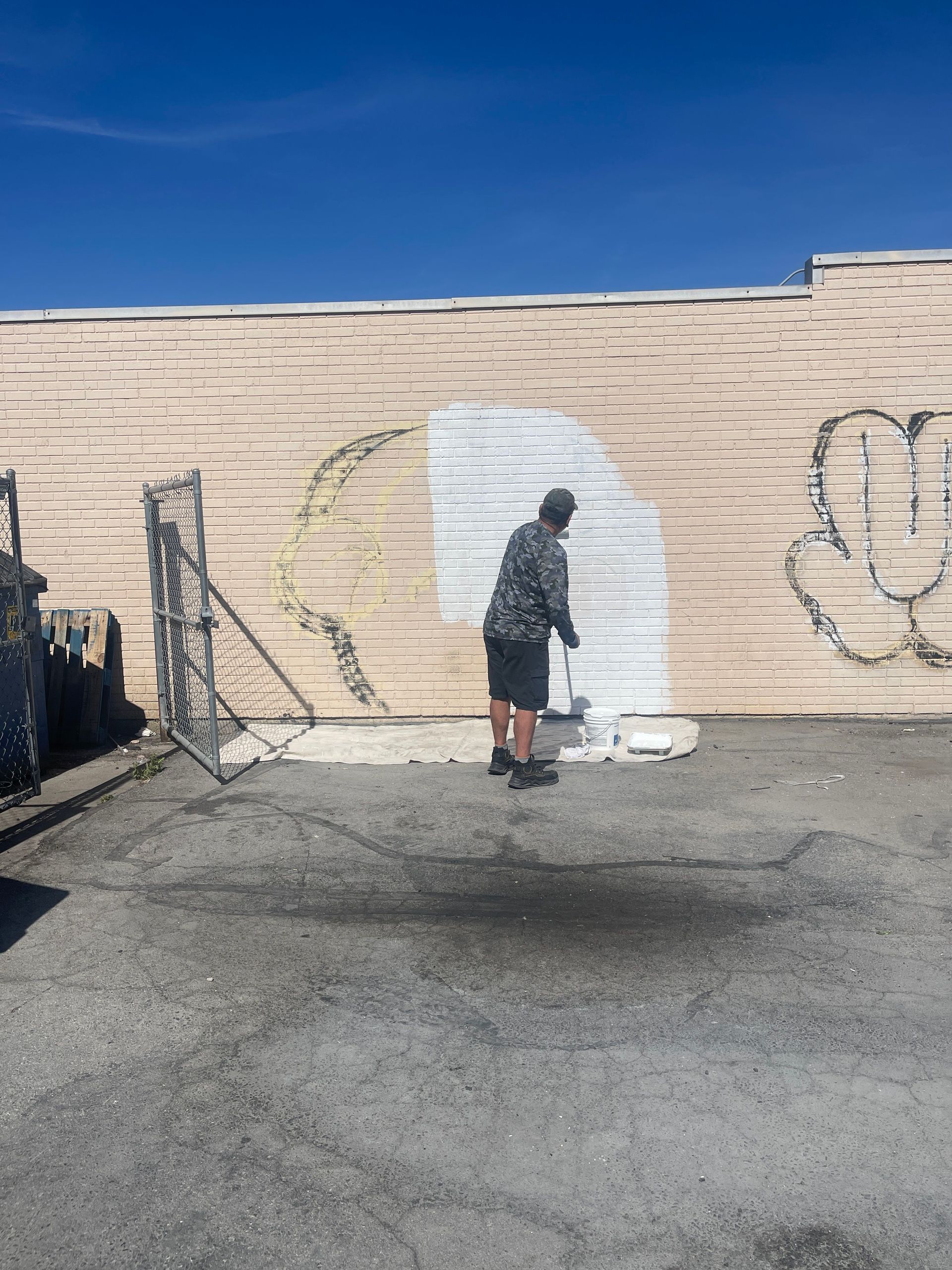 Person spraying white paint on a beige wall with graffiti in a sunny parking lot.