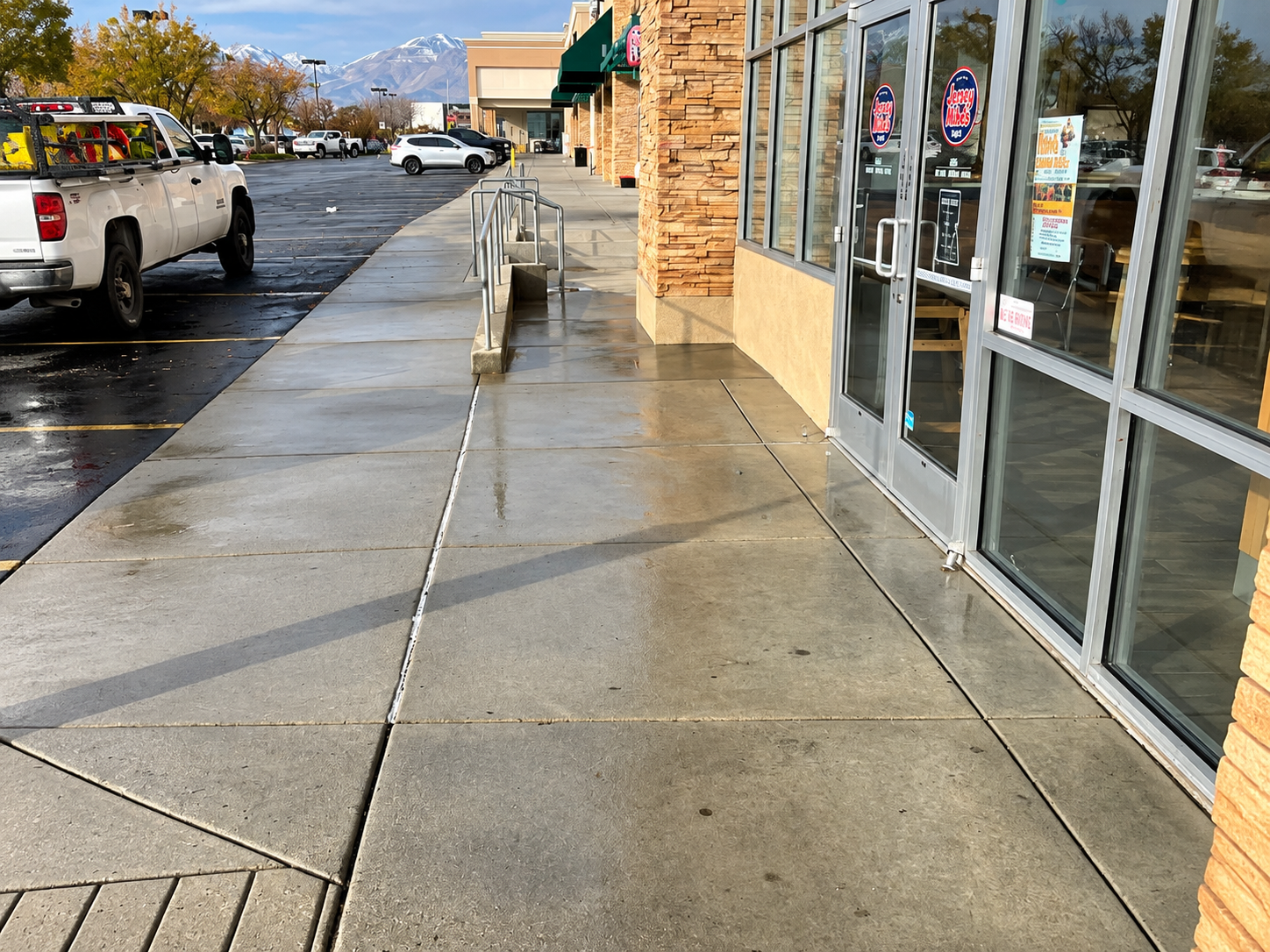 Wet sidewalk outside storefronts with parked trucks on a rainy street