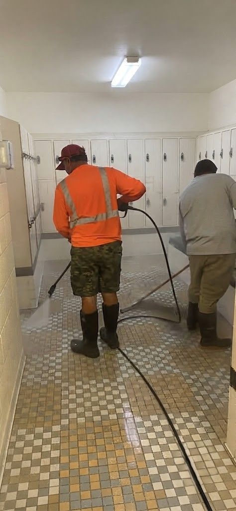Two workers in reflective vests are cleaning a tiled floor with hoses in a hallway.