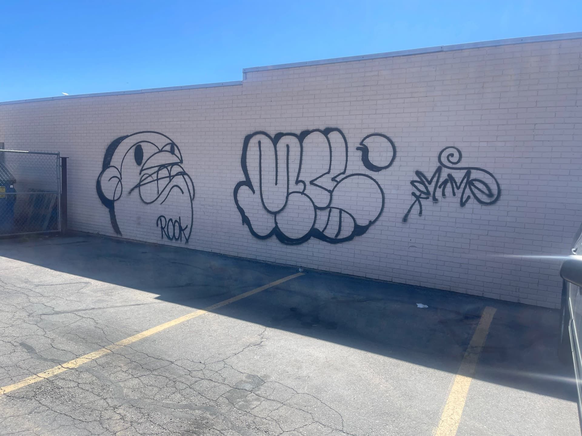 Graffiti tags on a white brick wall in a sunlit parking lot, with a sad face and a cartoon hand.