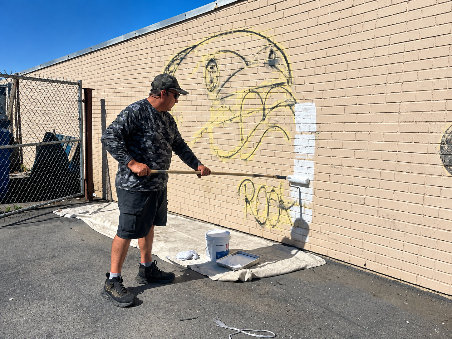 Person spray-painting graffiti on a beige brick wall beside a chain-link fence and paint bucket