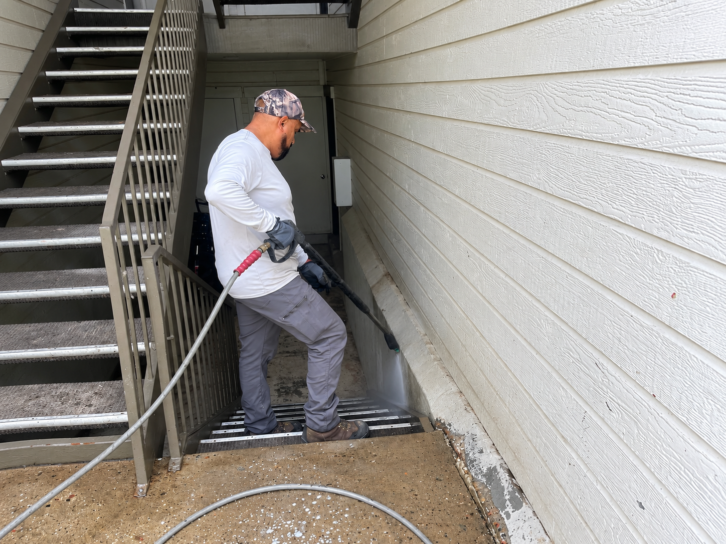 Worker pressure-washing exterior wall beside outdoor stairway, using hose and sprayer.