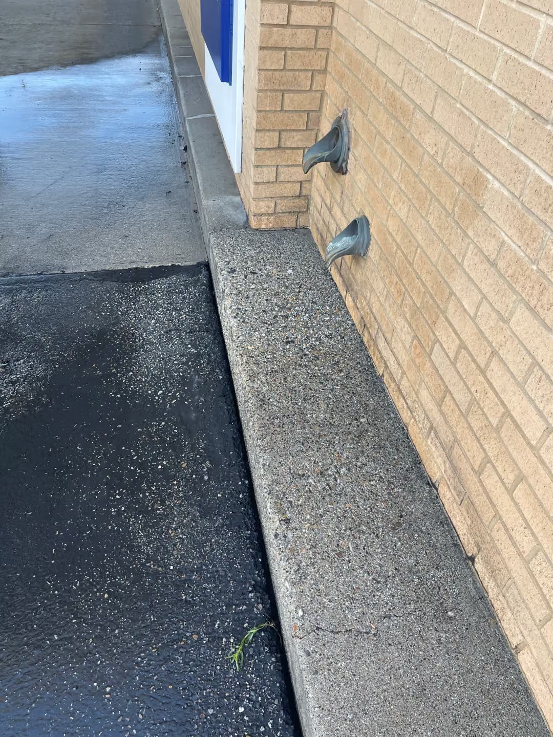 Brick wall beside asphalt walkway with two metal vents and a gravel strip along the base.