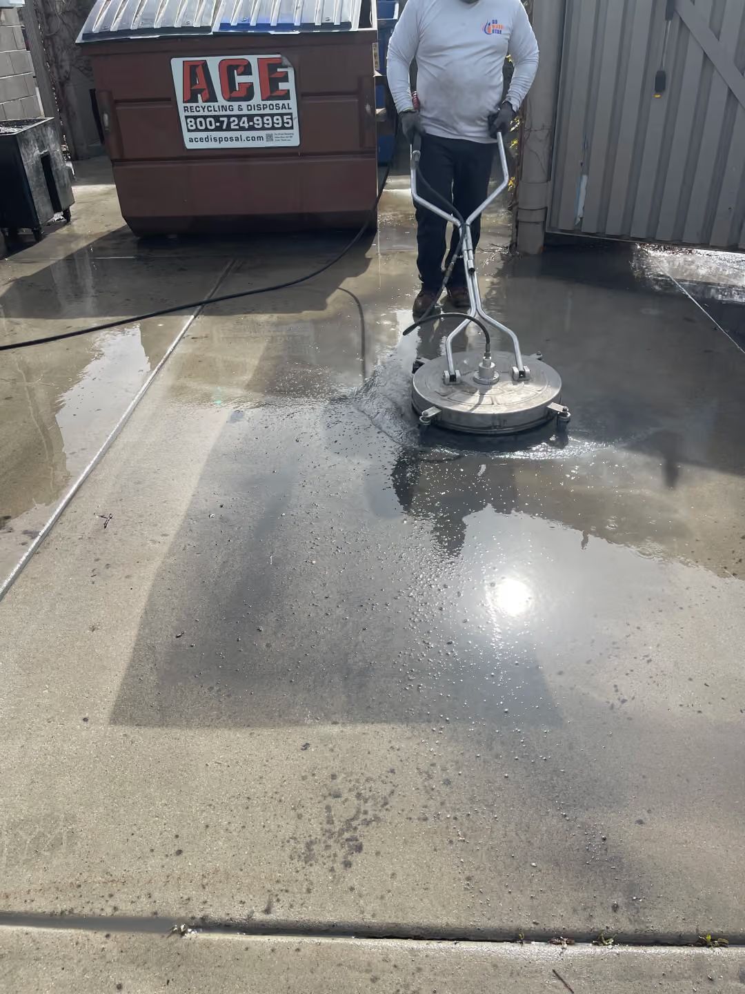 Person pressure washing a wet concrete surface beside a dumpster and parked vehicle.