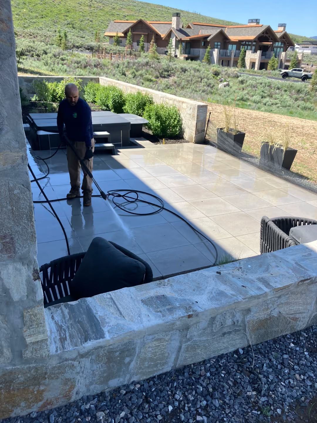Person pressure-washing a stone patio outdoors near a house and hillside.