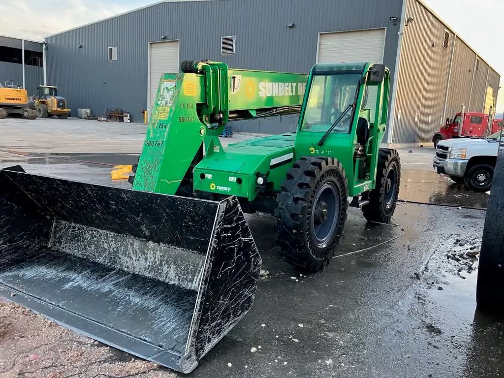 Green telehandler with front bucket parked outside a warehouse on a wet lot