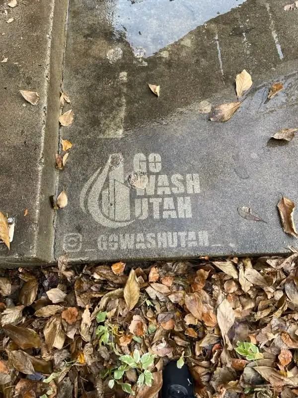 Storm drain cover with “Go Wash Utah” stencil, surrounded by wet leaves and dirt.