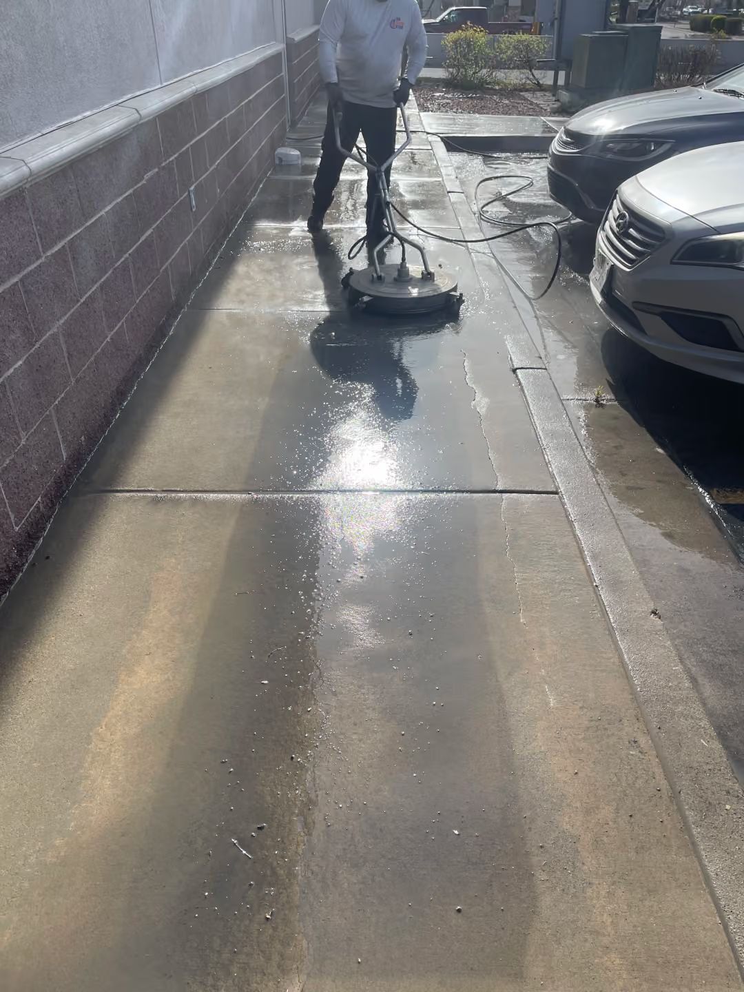 Person pressure-washing a wet sidewalk beside parked cars and a brick wall