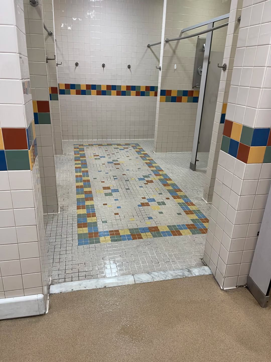 Public restroom shower stall with white and multicolor tile border and a tiled floor.